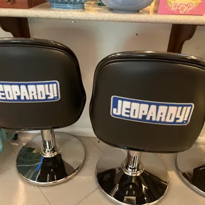 Sale Photo Thumbnail #57: Set of Jeopardy!-branded swivel bar stools featuring black vinyl upholstery, embroidered back logos, and chrome pedestal bases.