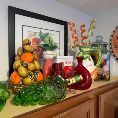 Sale Photo Thumbnail #36: Eclectic kitchen and dining decor display, including ceramic fruit topiary sculptures, green glass grape clusters, red art glass vases, and framed artwork.