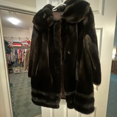 Sale Photo Thumbnail #130: Vintage dark brown faux fur coat featuring a wide shawl collar, wide horizontal bottom panel detail, and fully lined interior, hanging from a doorway.