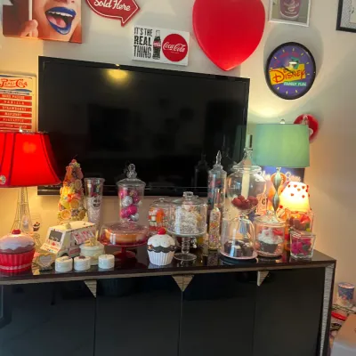 Sale Photo Thumbnail #7: Black lacquer media credenza topped with an array of colorful novelty decor, apothecary jars, lamps, and a flat-screen TV, framed by retro candy and soda signage on the wall above.