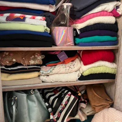 Sale Photo Thumbnail #173: Shelves filled with neatly folded women's clothing, including colorful sweaters, long-sleeve tops, and an assortment of handbags.