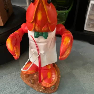 Sale Photo Thumbnail #223: Novelty anthropomorphic lobster waiter figure wearing a white apron and green bow tie, resting on a seashell-adorned base.