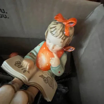 Sale Photo Thumbnail #98: Vintage Goebel Hummel porcelain figurine depicting a young girl with an orange hair bow reading a book, resting inside a cardboard box.
