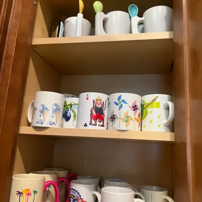 Sale Photo Thumbnail #29: Collection of assorted novelty and souvenir ceramic coffee mugs displayed on wood cabinet shelves, including casino-themed and colorful graphic designs.
