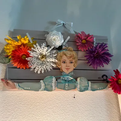 Sale Photo Thumbnail #120: Handcrafted wooden wall hanging featuring a painted Victorian-style lady cutout surrounded by colorful faux flowers on a slatted panel.