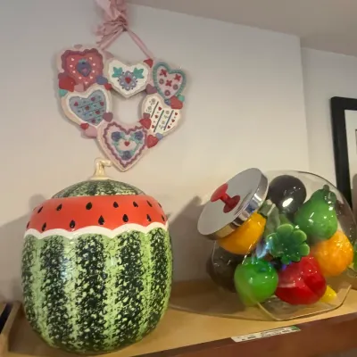 Sale Photo Thumbnail #34: Vintage ceramic watermelon cookie jar alongside a glass jar filled with colorful glass fruit decor, displayed beneath a heart-themed ceramic wall hanging.