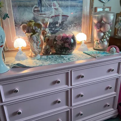 Sale Photo Thumbnail #189: Pink six-drawer dresser displayed with decorative figurines, a floral fishbowl, a glass jar filled with ornament balls, and a framed boat painting above.