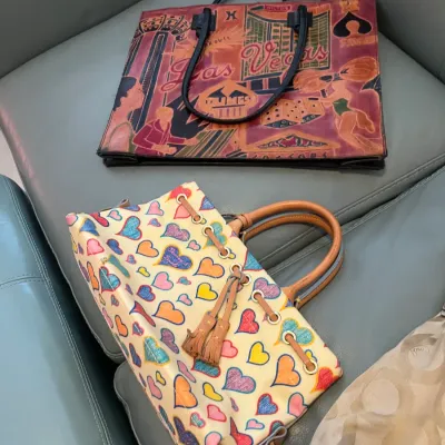 Sale Photo Thumbnail #48: Two leather handbags resting on a grey sofa, featuring a colorful Dooney & Bourke heart-pattern satchel with leather handles and a vintage embossed Las Vegas graphic tote bag.