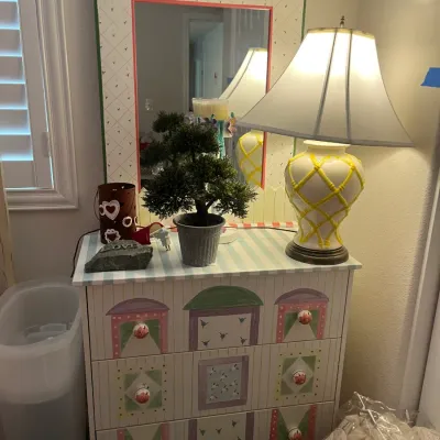 Sale Photo Thumbnail #240: Hand-painted dollhouse-style three-drawer dresser with matching house-framed vanity mirror, paired with a yellow latticework ceramic table lamp.