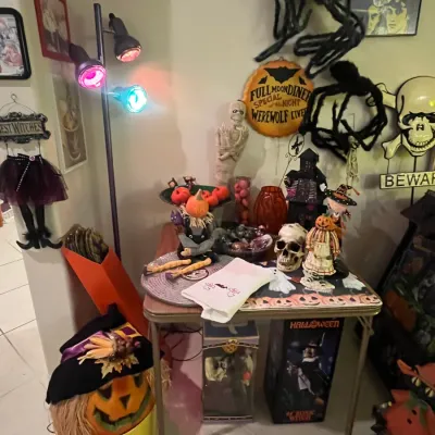 Sale Photo Thumbnail #72: Collection of Halloween decorations, featuring a floor lamp with colored bulbs, skull figures, a jack-o'-lantern, hanging spiders, and themed wall art.