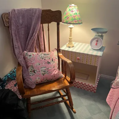 Sale Photo Thumbnail #241: Vintage carved oak wooden rocking chair draped with a plush blanket and pink throw pillow, set next to a hand-painted nightstand with a lamp and kitchen scale.