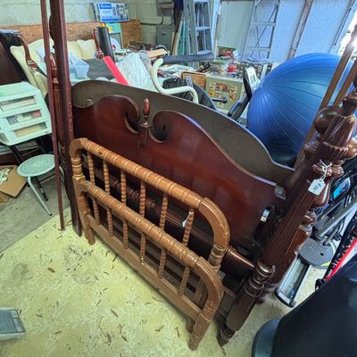 Sale Photo Thumbnail #12: Vintage mahogany four-poster bed frame featuring carved finials and decorative scrollwork, stored alongside a small wooden spindle-style safety rail in a workshop setting.