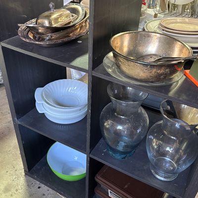 Sale Photo Thumbnail #62: Black wooden cube shelving unit displaying various household goods, including tarnished metal serving pieces, white ceramic bowls, glass vases, and a green Pyrex mixing bowl.