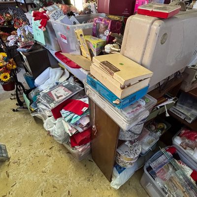Sale Photo Thumbnail #19: Crowded estate sale room featuring vintage sewing supplies, boxed games, craft materials, and plastic storage totes filled with various household goods.