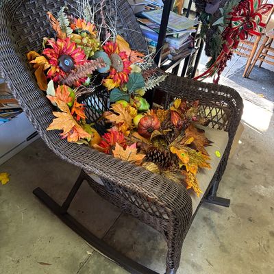 Sale Photo Thumbnail #37: Dark brown woven resin wicker rocking chair with a tan seat cushion, filled with an assortment of artificial autumn floral arrangements and foliage.
