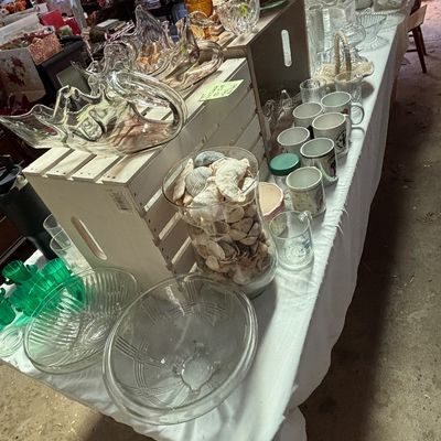 Sale Photo Thumbnail #57: Long table displayed with various glassware, including clear glass bowls, swan-shaped dishes, coffee mugs, and a large jar filled with seashells.