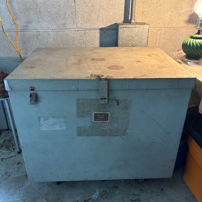 Sale Photo Thumbnail #109: Vintage heavy-duty military or industrial metal storage trunk with top latches and side metal plaque, showing surface wear and age.