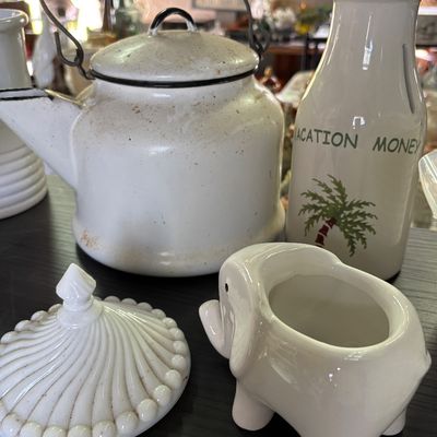 Sale Photo Thumbnail #63: Collection of white kitchen decor, featuring a vintage enamelware teapot with black rim, a ceramic elephant creamer, a milk bottle-style money jar, and a milk glass lid.