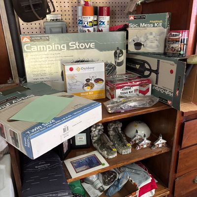 Sale Photo Thumbnail #45: Shelves filled with various outdoor gear including an Ozark Trail camping stove, mess kits, and a vinyl airbed, alongside decorative knick-knacks and household supplies.