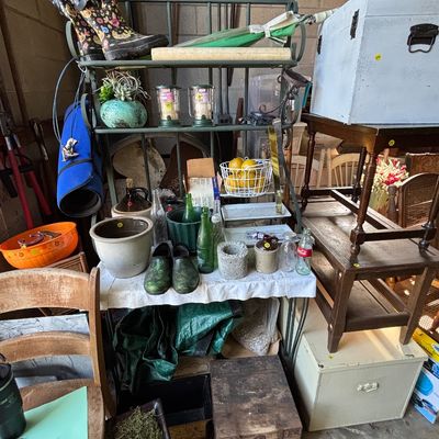 Sale Photo Thumbnail #29: A metal baker's rack holding various household goods, ceramic pottery, vintage bottles, rain boots, and lanterns, alongside stacked wooden tables and a rustic trunk.