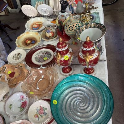 Sale Photo Thumbnail #65: Assorted collection of vintage glassware, ceramics, and tableware displayed on a white cloth, including a large teal swirl glass platter, red and gold urns, and decorative porcelain bowls.