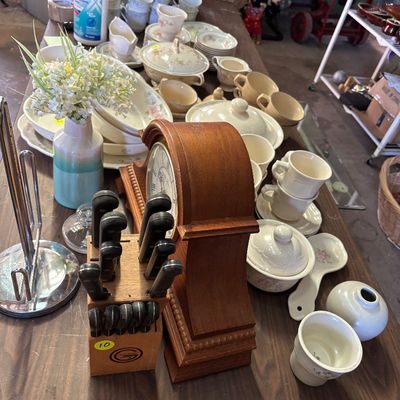 Sale Photo Thumbnail #73: A wooden mantle clock displayed alongside a knife block, ceramic dishware, teacups, and a small floral arrangement on a folding table.
