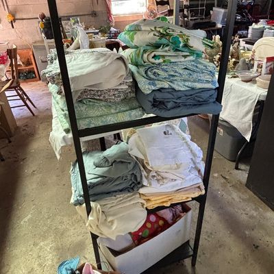 Sale Photo Thumbnail #52: Black metal shelving unit loaded with stacks of vintage bed linens, sheets, and blankets, along with a box of plush toys on the floor below.