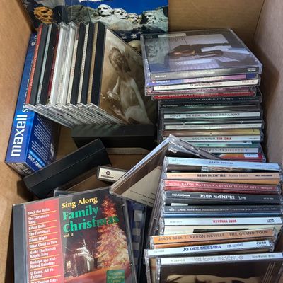 Sale Photo Thumbnail #13: Cardboard box packed with music CDs in jewel cases, VHS tapes, blank media boxes, and a Presidential Dollars collector folder.
