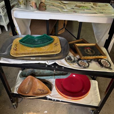 Sale Photo Thumbnail #67: A tiered utility cart displaying various home goods, including ceramic trays, framed artwork, wooden bowls, and colored dinnerware plates.
