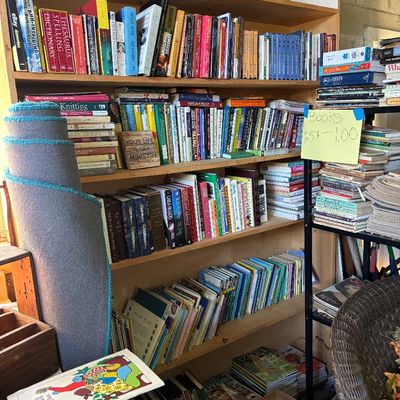 Sale Photo Thumbnail #38: Large wooden bookcase filled with various hardcover and paperback books, alongside a metal shelf unit holding additional stacks of books priced from $0.25 to $1.00.