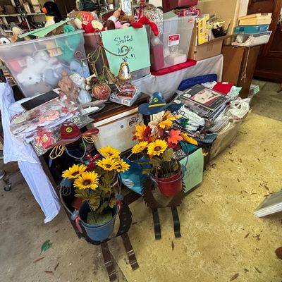 Sale Photo Thumbnail #21: Collection of sale items including metal rooster plant holders with artificial sunflowers, plastic storage bins of plush toys, and craft supplies in a garage setup.