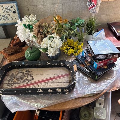 Sale Photo Thumbnail #69: A collection of household goods featuring a rustic wooden serving tray with a Chianti graphic, Star Wars puzzles, artificial floral arrangements, and a small green glass lamp.