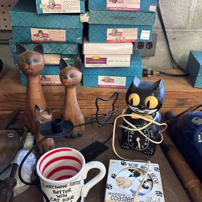 Sale Photo Thumbnail #79: Collection of cat-themed decor including carved wooden cat figurines, a novelty cat clock, a coffee mug, and absorbstone coasters, with vintage Madame Alexander doll boxes stacked behind.