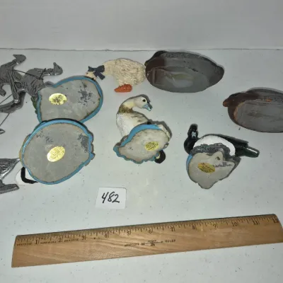 Sale Photo Thumbnail #828: Collection of small waterfowl figurines, duck decoys, and metal bird decorations, displayed with a wooden ruler for scale.