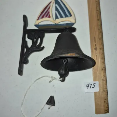 Sale Photo Thumbnail #817: Cast iron wall-mounted dinner bell with a painted sailboat topper and decorative scrollwork bracket, shown alongside a wooden ruler for scale.