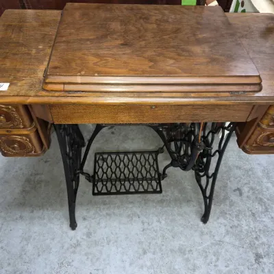 Sale Photo Thumbnail #187: Antique treadle sewing machine in a wooden cabinet featuring carved drawers, a hinged top cover, and a black cast iron base.