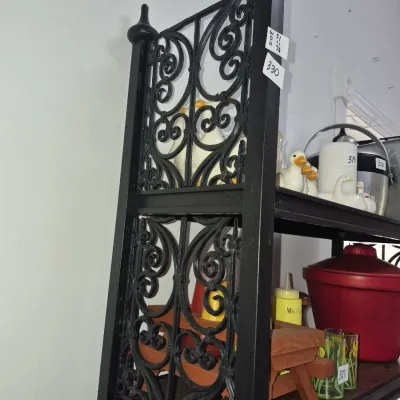 Sale Photo Thumbnail #576: Black wrought iron bakers rack featuring ornate scrollwork sides and decorative finials, holding various kitchenware and collectibles.