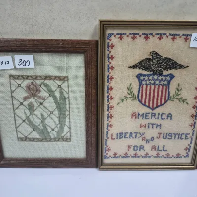 Sale Photo Thumbnail #534: Two framed needlepoint art pieces, featuring a floral motif in a dark wood frame and an American patriotic emblem with text in a metallic gold-tone frame.
