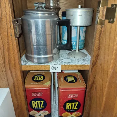 Sale Photo Thumbnail #723: Vintage aluminum stovetop percolator with glass knob, shown alongside two Ritz Crackers limited edition collectible tins and other kitchenware stored in a cabinet.