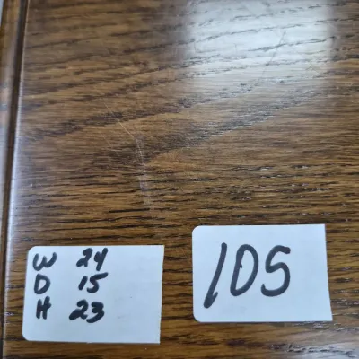 Sale Photo Thumbnail #171: Close-up view of a wooden furniture surface displaying lot tag 105 and handwritten dimensions of 24 inches wide, 15 inches deep, and 23 inches high.
