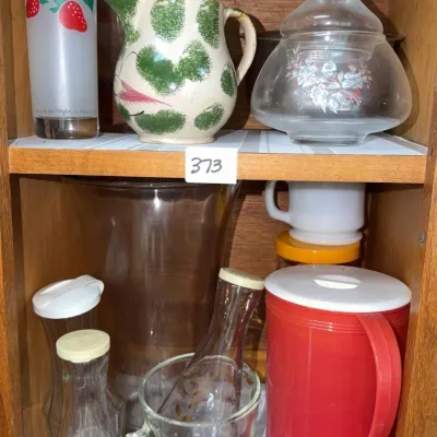 Sale Photo Thumbnail #647: Wooden cabinet shelves filled with assorted glassware and kitchenware, including a vintage strawberry drinking glass, a ceramic pitcher, and a red plastic pitcher.