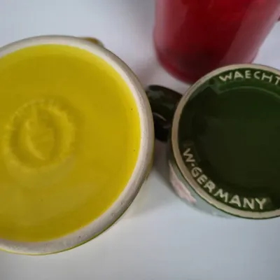 Sale Photo Thumbnail #609: Underside of colorful vintage ceramic mugs, including a green Waechtersbach West Germany mug and a yellow ceramic mug, showcasing manufacturer backstamps.