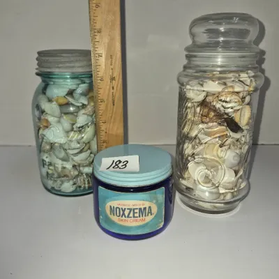 Sale Photo Thumbnail #315: Collection of vintage glass jars filled with seashells, including a blue Ball Mason jar, a tall clear glass canister, and a vintage cobalt blue Noxzema skin cream jar.