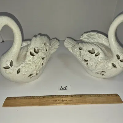 Sale Photo Thumbnail #229: Pair of off-white ceramic swan figurines featuring intricate reticulated or cutout wing details, displayed alongside a wooden ruler for scale.