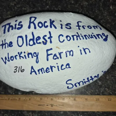 Sale Photo Thumbnail #552: Hand-painted commemorative stone bearing the inscription 'This Rock is from the Oldest Continuing Working Farm in America', displayed next to a ruler for scale.