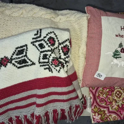 Sale Photo Thumbnail #80: Collection of vintage home textiles including a cream cable knit blanket, a cross-stitched throw with red and black geometric patterns, and a country-style throw pillow featuring an embroidered winter scene.