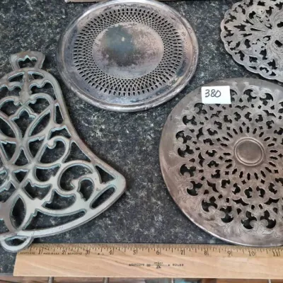 Sale Photo Thumbnail #660: Collection of four vintage metal trivets and coasters, including a bell-shaped cast trivet and pierced silver-plated pieces with age-appropriate patina.