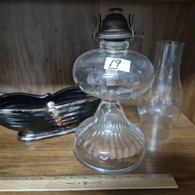 Sale Photo Thumbnail #30: Vintage clear pressed glass kerosene oil lamp with a ribbed pedestal base, brass burner, and separate glass chimney, displayed alongside a black ceramic boat-shaped planter.