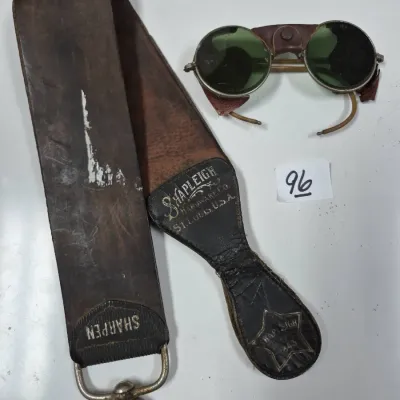 Sale Photo Thumbnail #152: Vintage Shapleigh Hardware Co. leather razor strop with metal swivel ring, paired with early 20th-century leather side-shield safety goggles with dark green lenses.