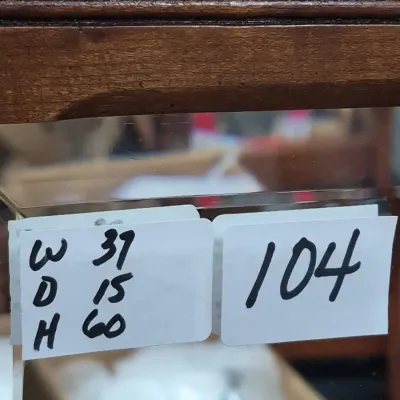 Sale Photo Thumbnail #168: Handwritten tag displaying dimensions and lot number 104 affixed to a wooden display cabinet or piece of furniture.